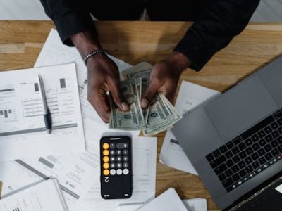 Person counting US dollars, using a calculator and laptop, with financial documents on a wooden desk.